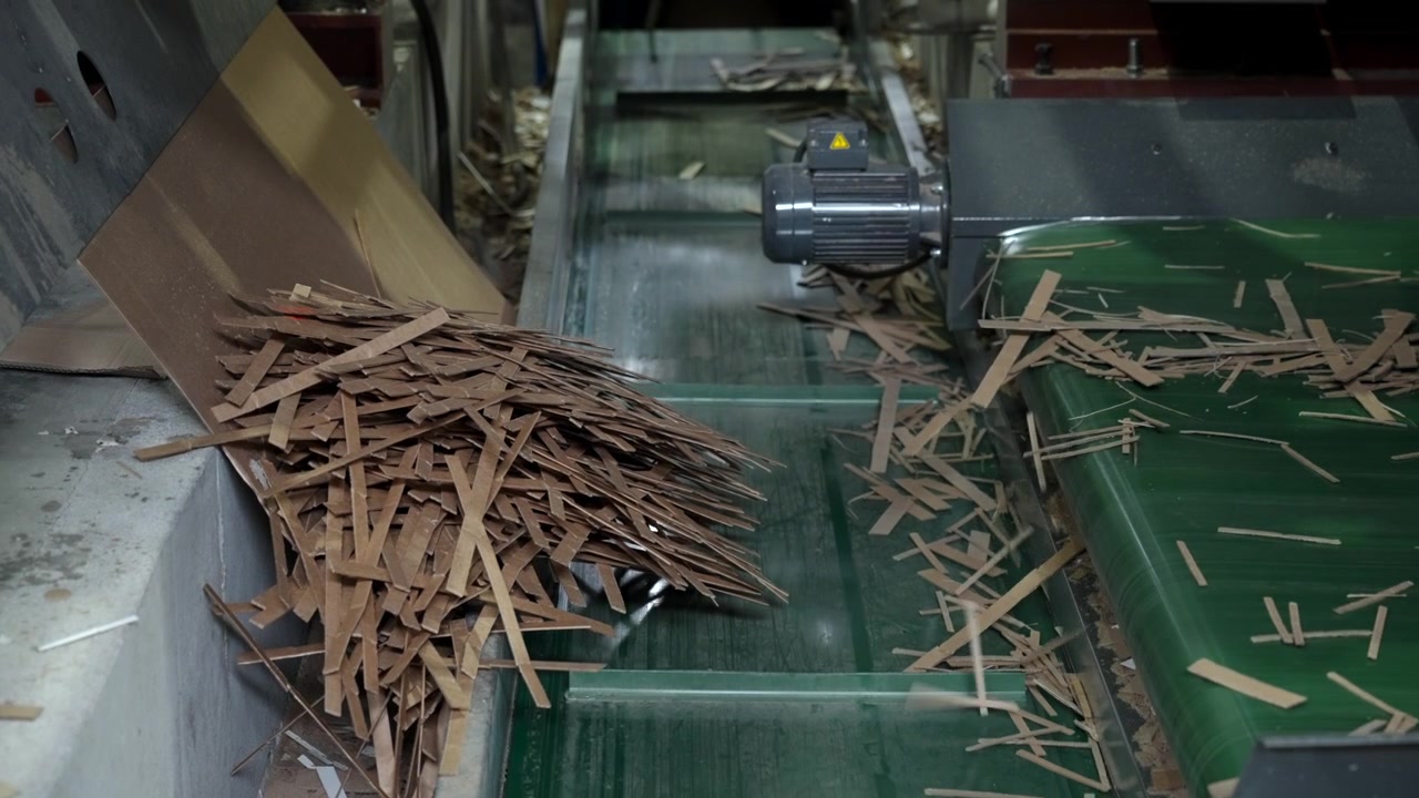 Close view of factory processing line showing stacked cardboard strips on left chute, loose scrap across green conveyor surface, central motor housing, and worn metal machinery under dim indoor light