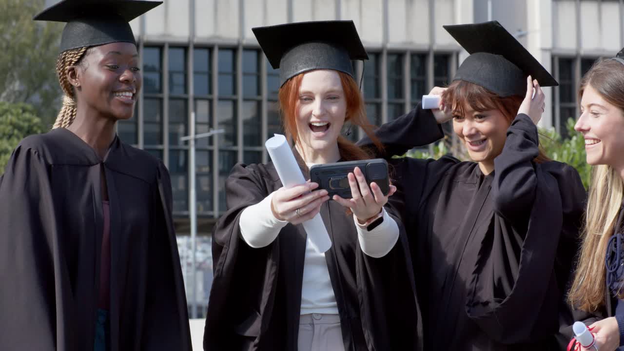 En rødhåret dimittend løfter sin smartphone for at tage en selfie, mens dimittender af forskellig baggrund stiller sig op og vifter med diplomer på campus.