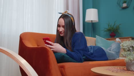 joven mujer caucásica acostada en el sofá usando teléfono inteligente viendo videos, compras en línea, tocando