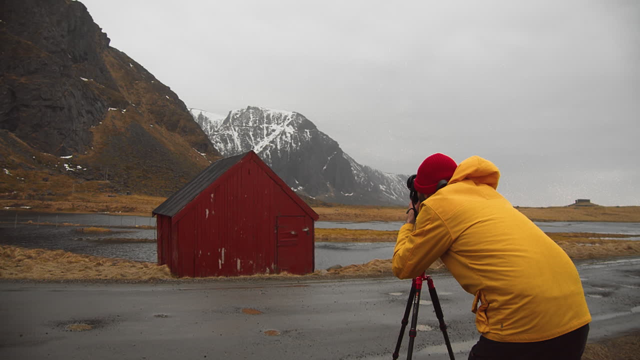 Photographer Wearing Yellow Hood Jacket And Red Cap Positioned In Front Of A Red House While Taking A Picture Of Mountains- Static
