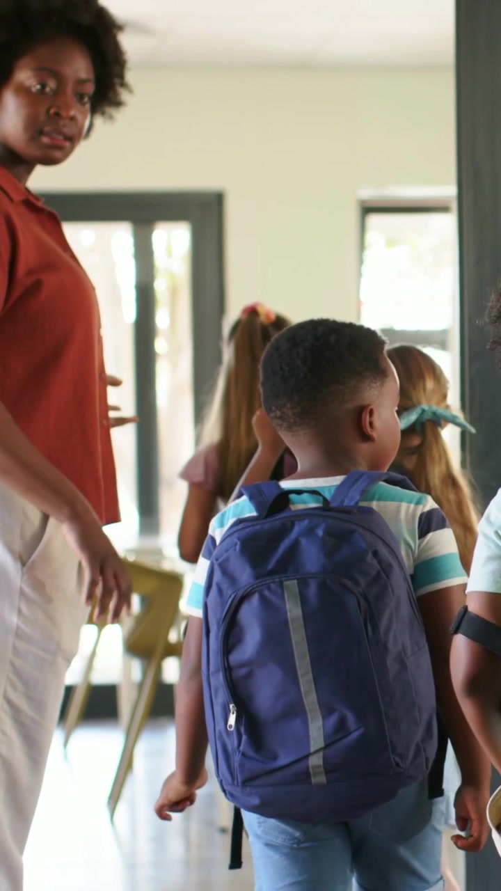 Vertical video: Walking kids following teacher in rust top gesturing into classroom with backpacks