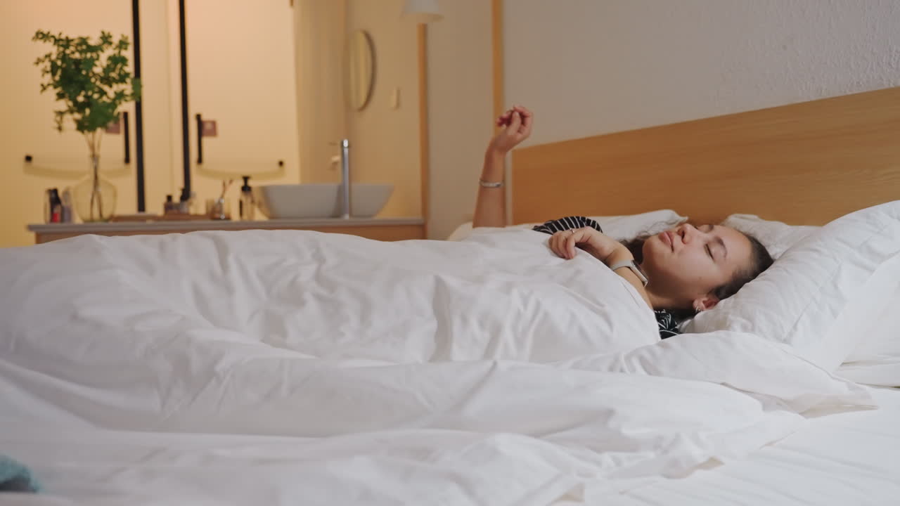 Kvinde vågner i hotelseng under dynen, nærbillede af gæst der putter sig i blødt hvidt sengetøj og pude på hyggeligt hotelværelse med varm sengelampe, lille plante på hylden, synlig vask og spejl i baggrunden.