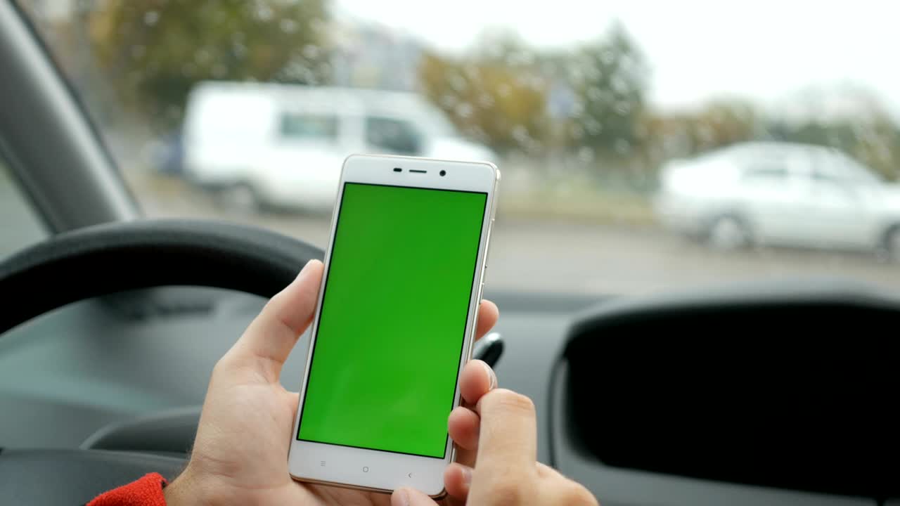 mano sosteniendo teléfono inteligente con pantalla verde en blanco en el coche para la dirección, masaje, ubicación, negocio. vida de la ciudad. hombre se sienta en el coche y trabaja en el teléfono inteligente pantalla verde de cerca. llave de croma. día lluvioso.