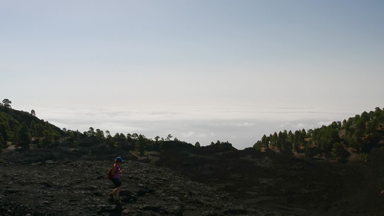 Drone shot following a woman hiking on top of a volcano. In the background there is a sea of clouds
