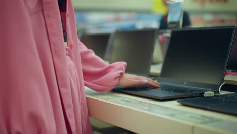 vista del cuerpo de la dama en tela rosada con bolso negro interactuando con una computadora portátil en exhibición en una tienda electrónica, la pantalla de la computadora portable está en blanco mientras la opera, con otras computadoras portátiles ordenadamente alineadas