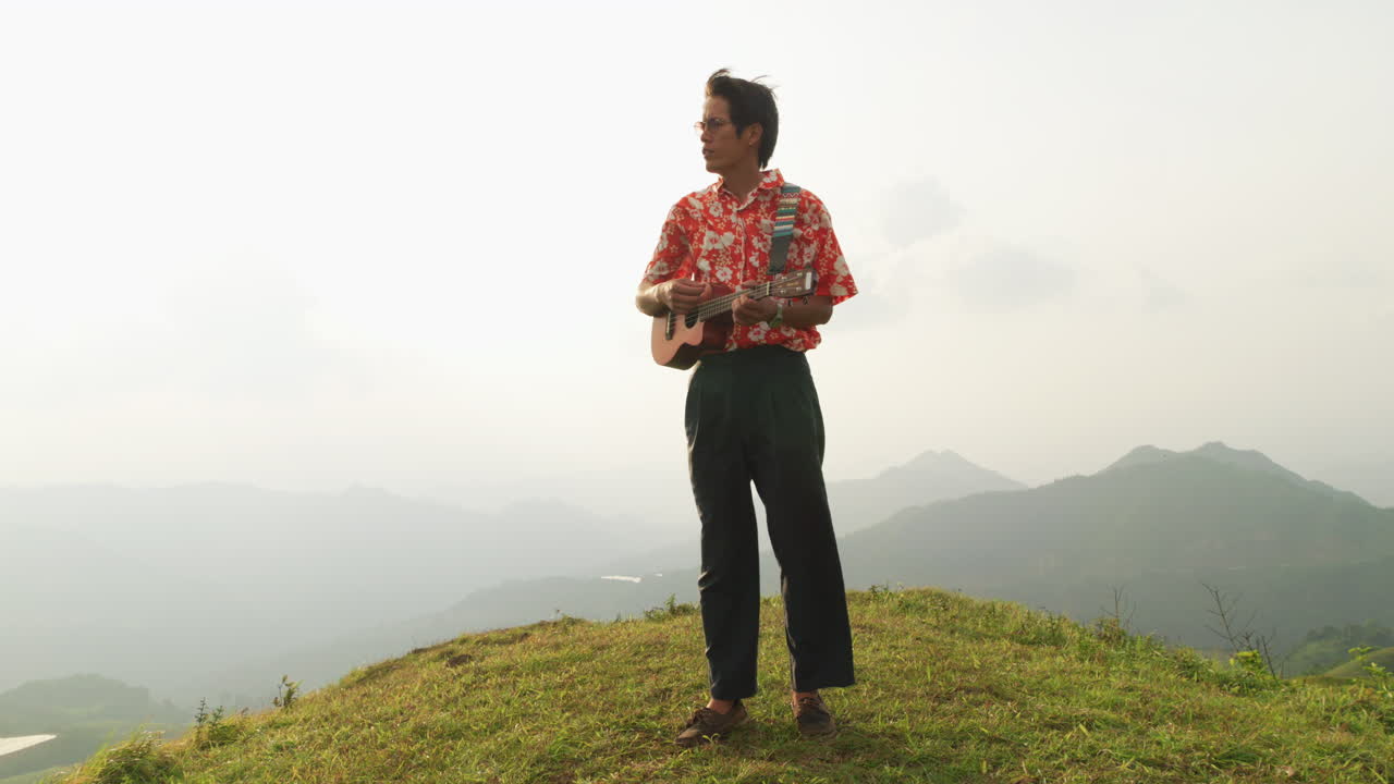 Static shot of an Asian man standing on top on a mountain singing while playing ukulele on a cloudy day