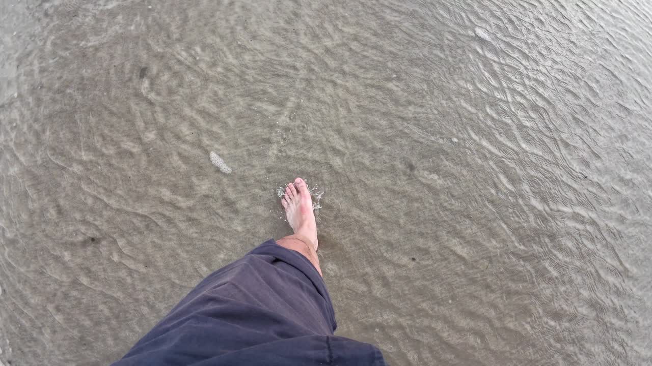 A first-person view of someone walking barefoot through shallow water on Polzeath Beach, UK, as gentle waves ripple over the sand and seaweed drifts nearby, capturing a calm seaside moment