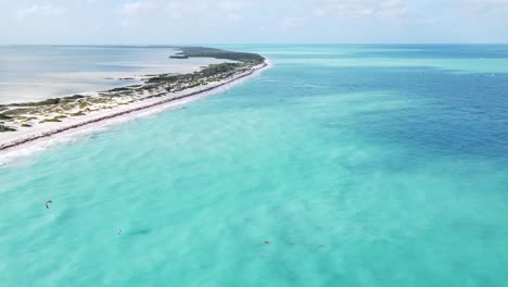 Drone-views-of-the-azure-and-turquoise-waters-of-the-Caribbean-Sea-in-Isla-Blanca,-Cancun,-Mexico,-while-a-kite-surfer-is-flying-over-the-pristine-blue-water-washing-over-the-white-beach