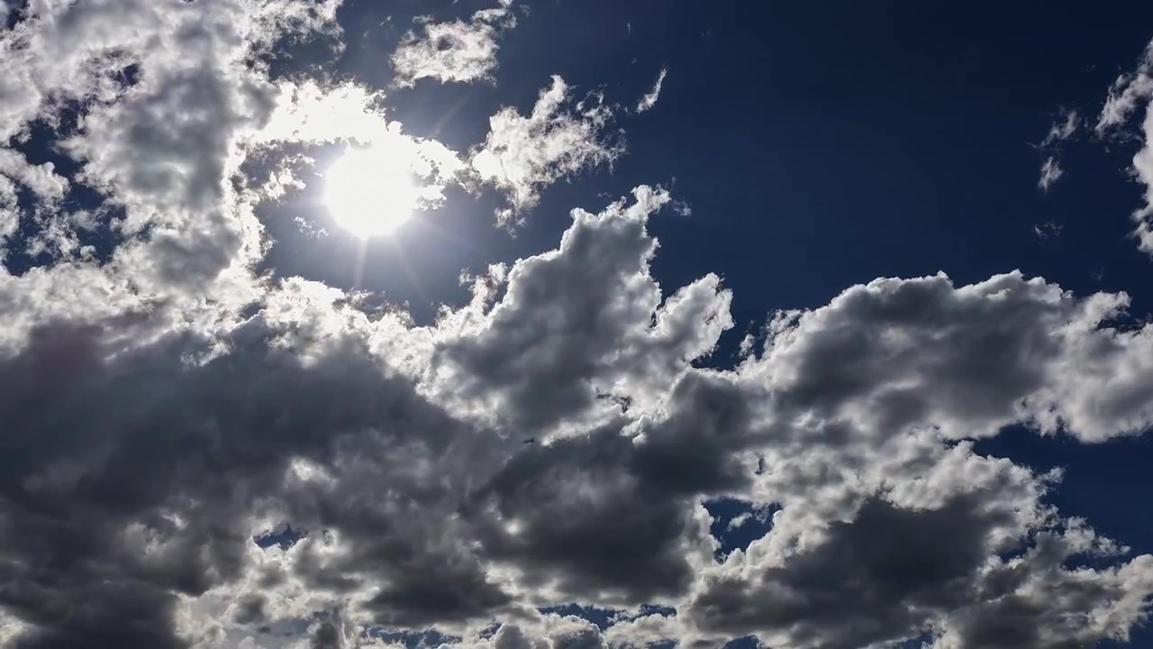 Timelapse estável 4K UHD de um céu azul profundo e vívido perfurado pelo sol brilhante. Nuvens prateadas e cinza-cromado em movimento rápido e altamente contrastadas se deslocam dinamicamente pela moldura ampla