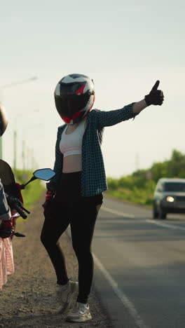 two women by the roadside, one seated on a motorcycle playing with her clothes while the other stands and gives a thumbs up, a car approaches in the distance