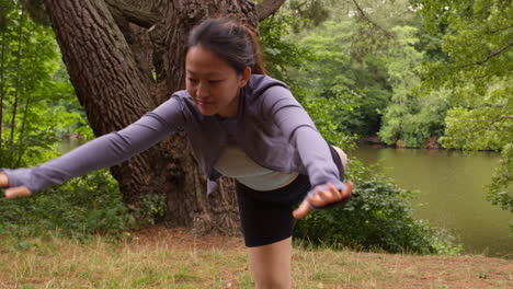 woman wearing sports clothing doing yoga stretching and balancing in forest by lake or river enjoying peace and beauty of nature