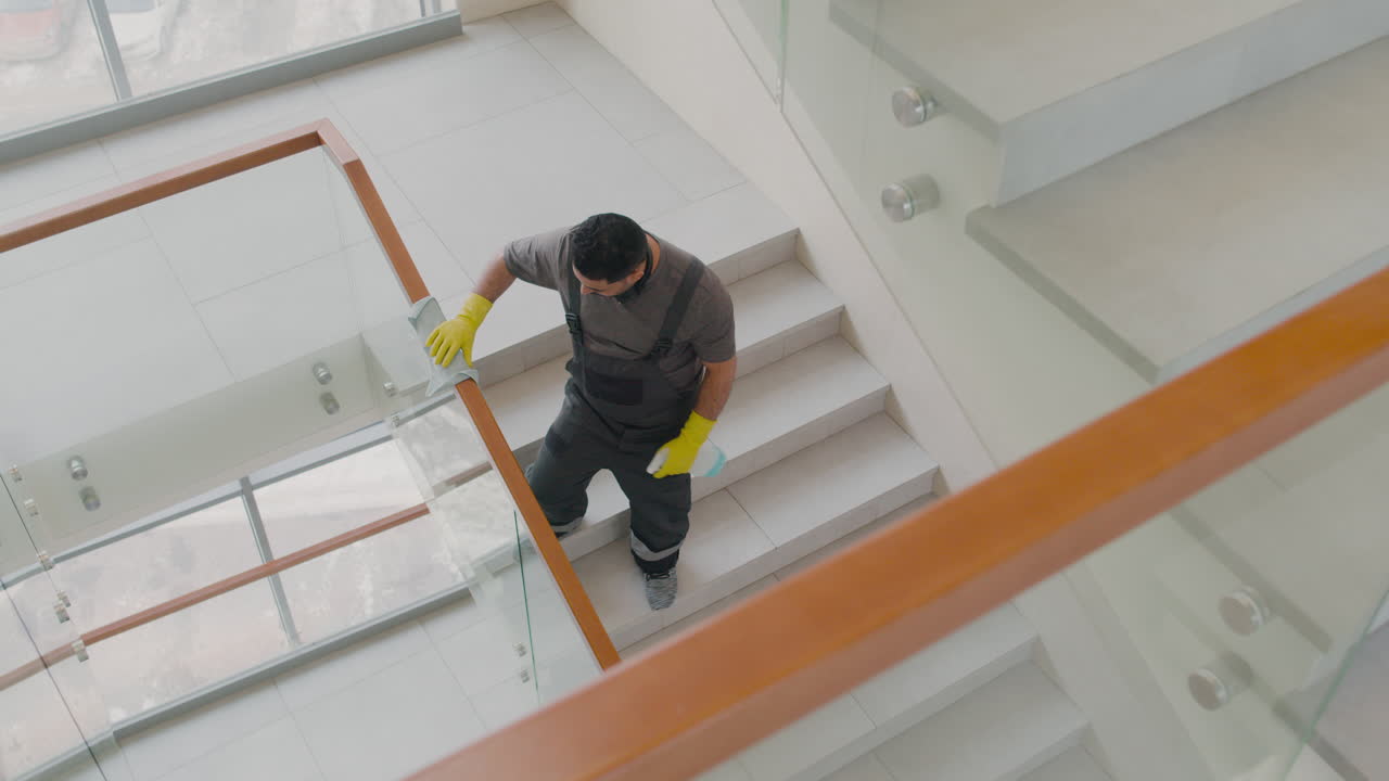 vista superior de un hombre de limpieza árabe con guantes limpiando barandillas de escaleras y cristales dentro de un edificio de oficinas