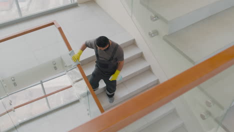 Blick-Von-Oben-Auf-Einen-Arabischen-Putzmann-Mit-Handschuhen,-Der-Treppengeländer-Und-Kristalle-In-Einem-Bürogebäude-Reinigt