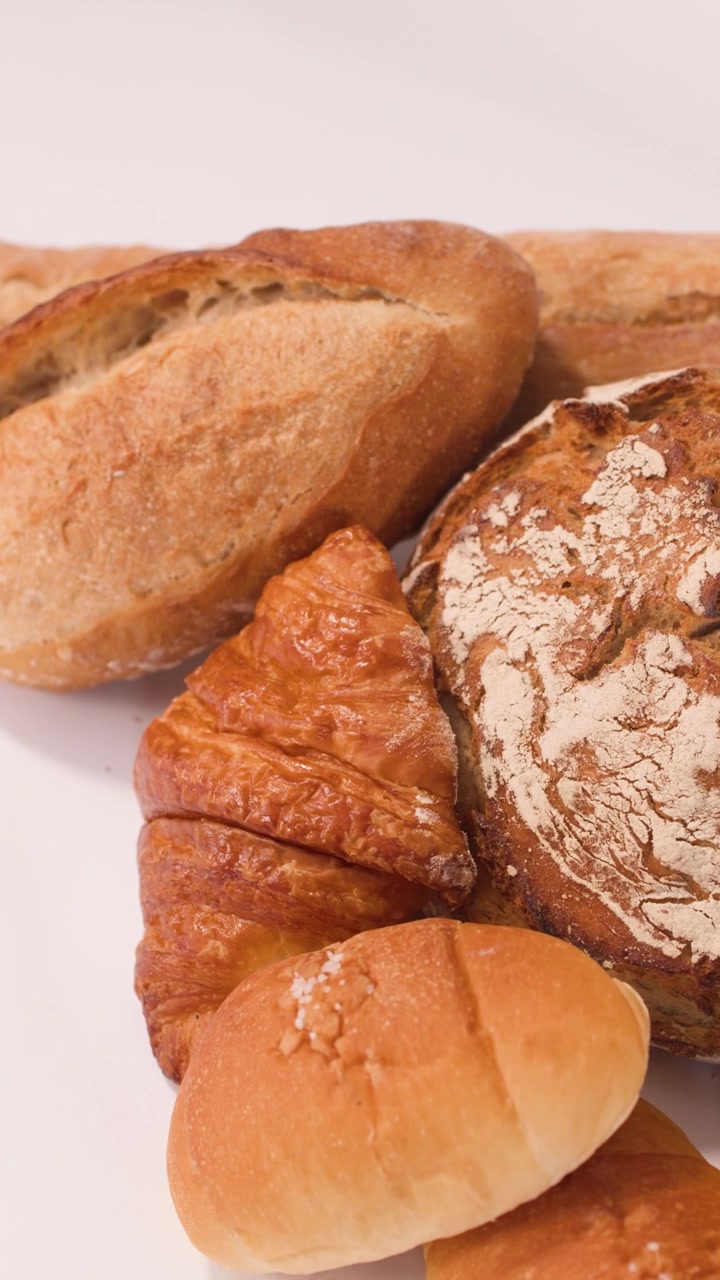Slow camera zoom over sourdough, French baguettes, croissants, and sliced grains on white surface.