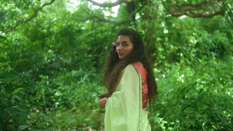 a woman dressed in a green sari and red top turns her back to the camera and walks into the forest