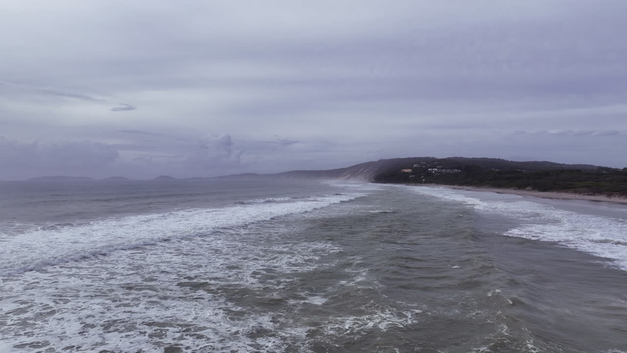 Flight over wild breaking waves on Rainbow Beach towards misty subdued bay