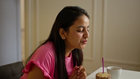vista lateral de una feliz chica india morena en un vestido rosa soplando una vela en un pequeño pastel y aplaudiendo sus manos durante una celebración de cumpleaños en un apartamento moderno durante el día