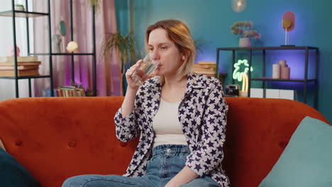 satisfied caucasian woman sitting at home on couch leading healthy lifestyle drinking pure water