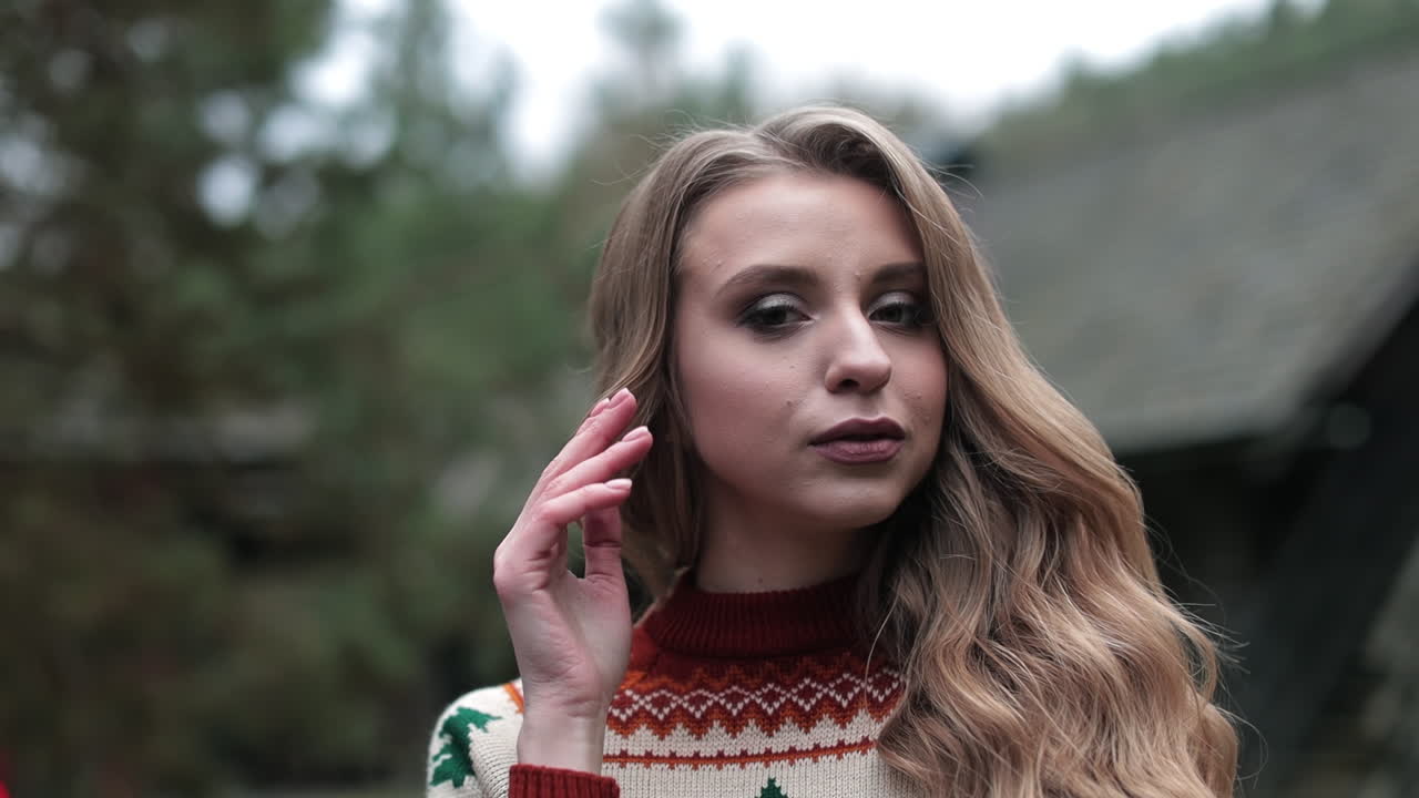 Fair-haired woman with bright makeup posing in front of the camera. Young female in warm sweater at the blurred background. Close up.