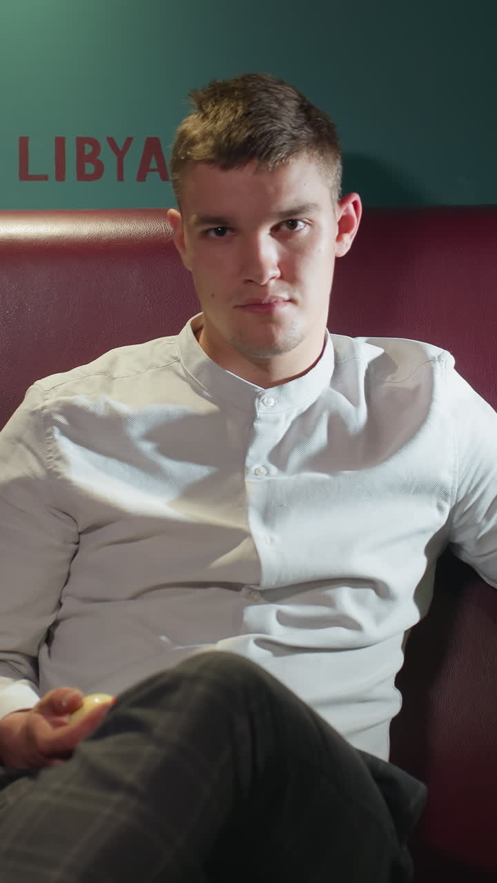 색 셔츠와 칠색 바지를 입은 젊은이가 빌리어드 방의 빨간 소파에 앉아 손에 오렌지색 빌리어드를 고 있다. 큐 스틱이 에 기대어 있다. 배경에는 녹색 벽에 나라 이름이 그려져 있다.