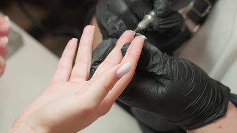 close up of nail technician wearing black gloves carefully filing inside of acrylic nail on client hand using electric drill tool. white polished nails and gentle grip highlight precision and care