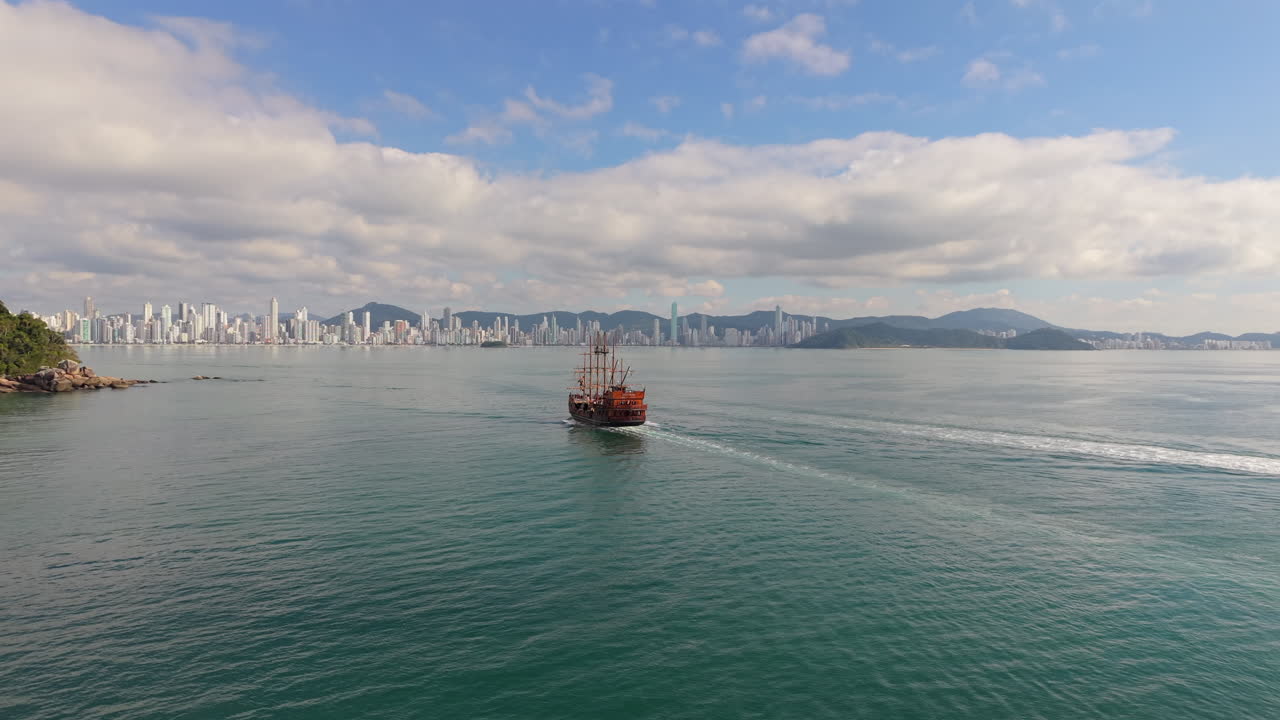 Following aerial of pirate ship sailing on ocean and moving towards Balneário Camboriú coastal city shoreline, Santa Catarina, Brazil