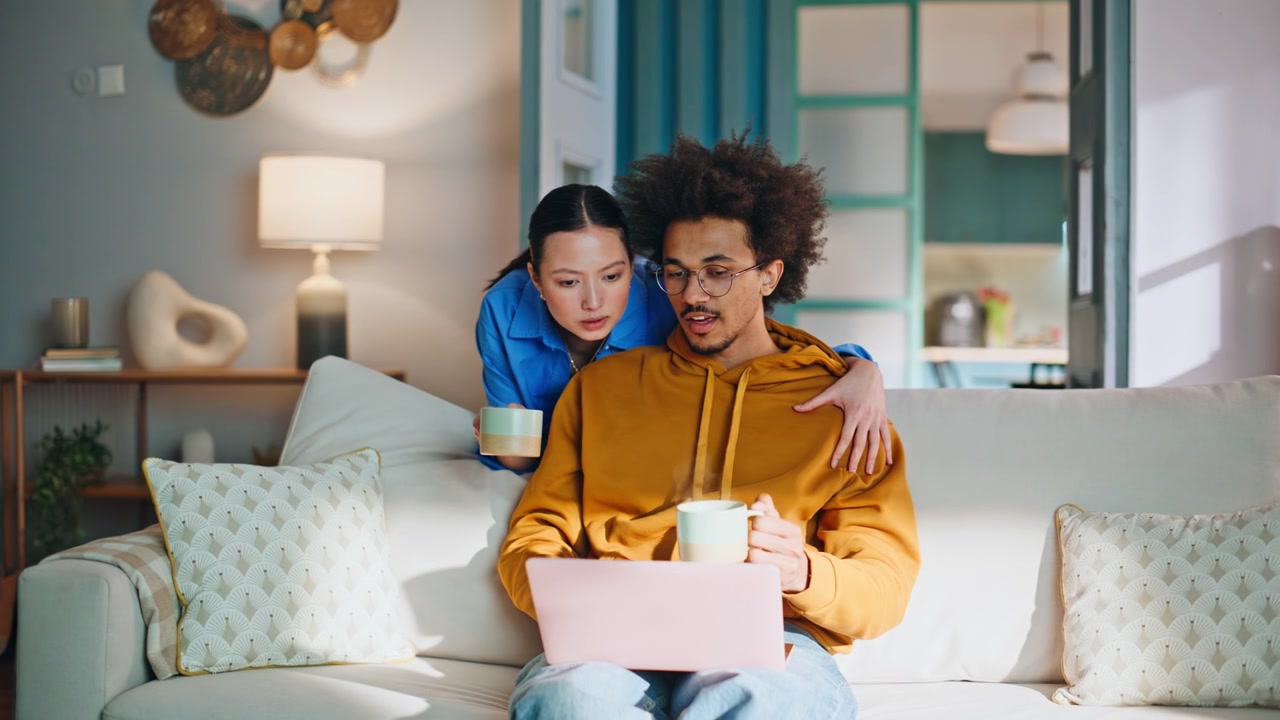Couple browsing laptop sofa home. Woman hugs boyfriend with coffee mugs.