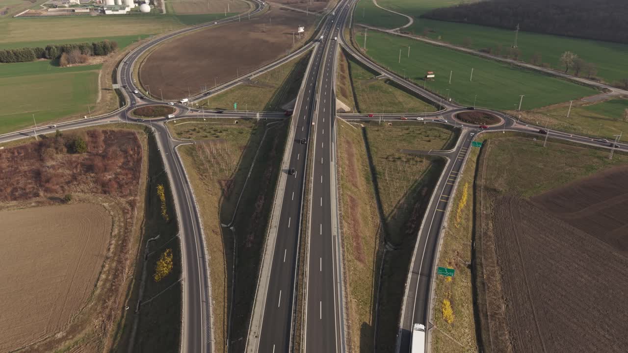 A high-angle aerial view of a highway interchange surrounded by agricultural land. Multiple roads, roundabouts, and connecting lanes form a structured transport network with visible vehicle traffic