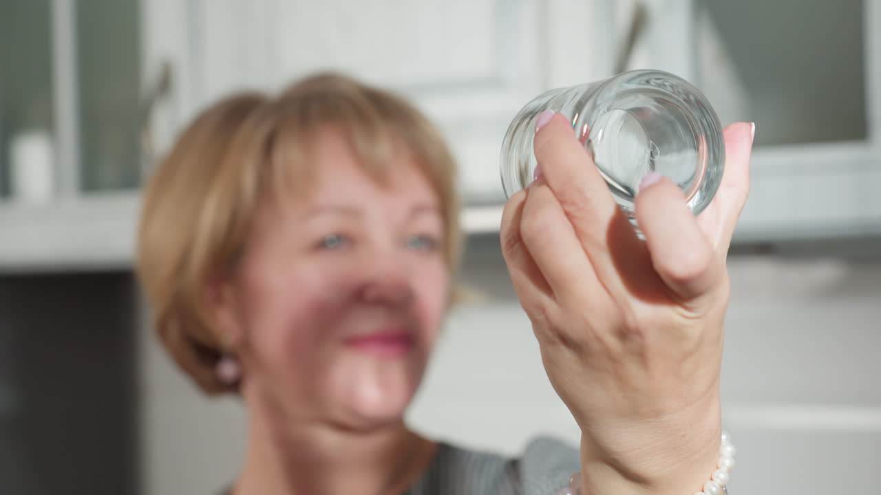 Lady slightly observing sparkling clean transparent glass cup with polished nails, holding it carefully under bright kitchen light in front of white modern cabinet