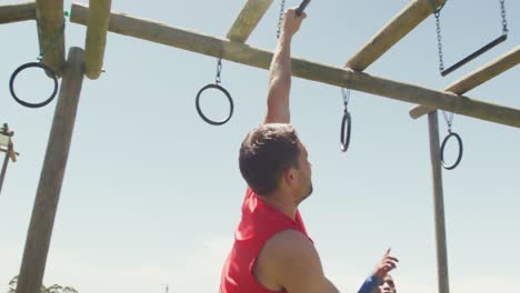 hombre caucásico en forma en chaleco rojo colgando de barras de trapecio en una carrera de obstáculos en el sol