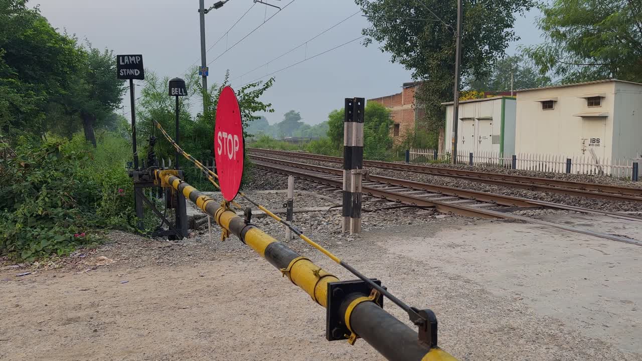 Slow push-in shot toward a railway crossing with a red stop sign, striped barrier, gravel ground, metal tracks, green vegetation, distant buildings, and a calm roadside atmosphere