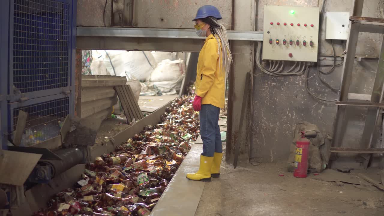 Woman-worker in yellow and transparent protecting glasses, hard hat and mask watching the conveyor full of used plastic bottles. Footage of automized process on recycle plant
