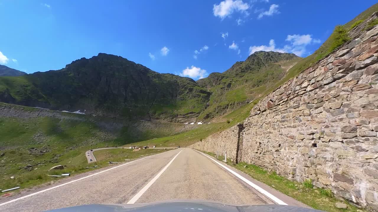 A Driver’s Perspective on Transfagarasan Mountain Highway, Surrounded by Towering Green Peaks with Patches of Snow and a Clear Blue Sky
