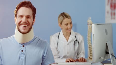 Paciente-Sonriendo-A-La-Cámara-Mientras-La-Fisioterapeuta-Trabaja-Sobre-La-Computadora-En-Segundo-Plano