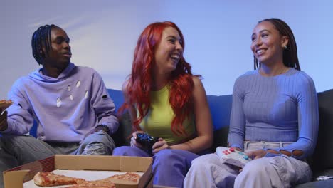 group of gen z friends sitting on sofa at home gaming and eating takeaway pizza 3