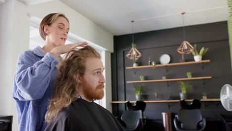 una feliz peluquera caucásica aconsejando a un cliente masculino con cabello largo en el salón, en cámara lenta