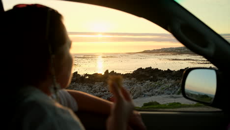 chica disfruta de la puesta de sol a través de la ventana del coche mientras come un bocadillo, vista de perfil