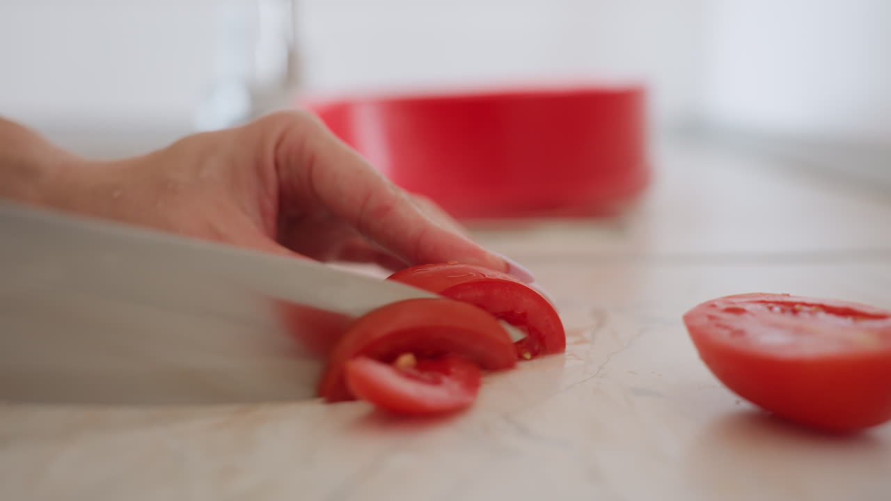 Close up of chef hand slicing fresh tomato with sharp knife on kitchen counter, preparing ingredient for healthy meal, highlighting detail of red vegetable and careful cooking process
