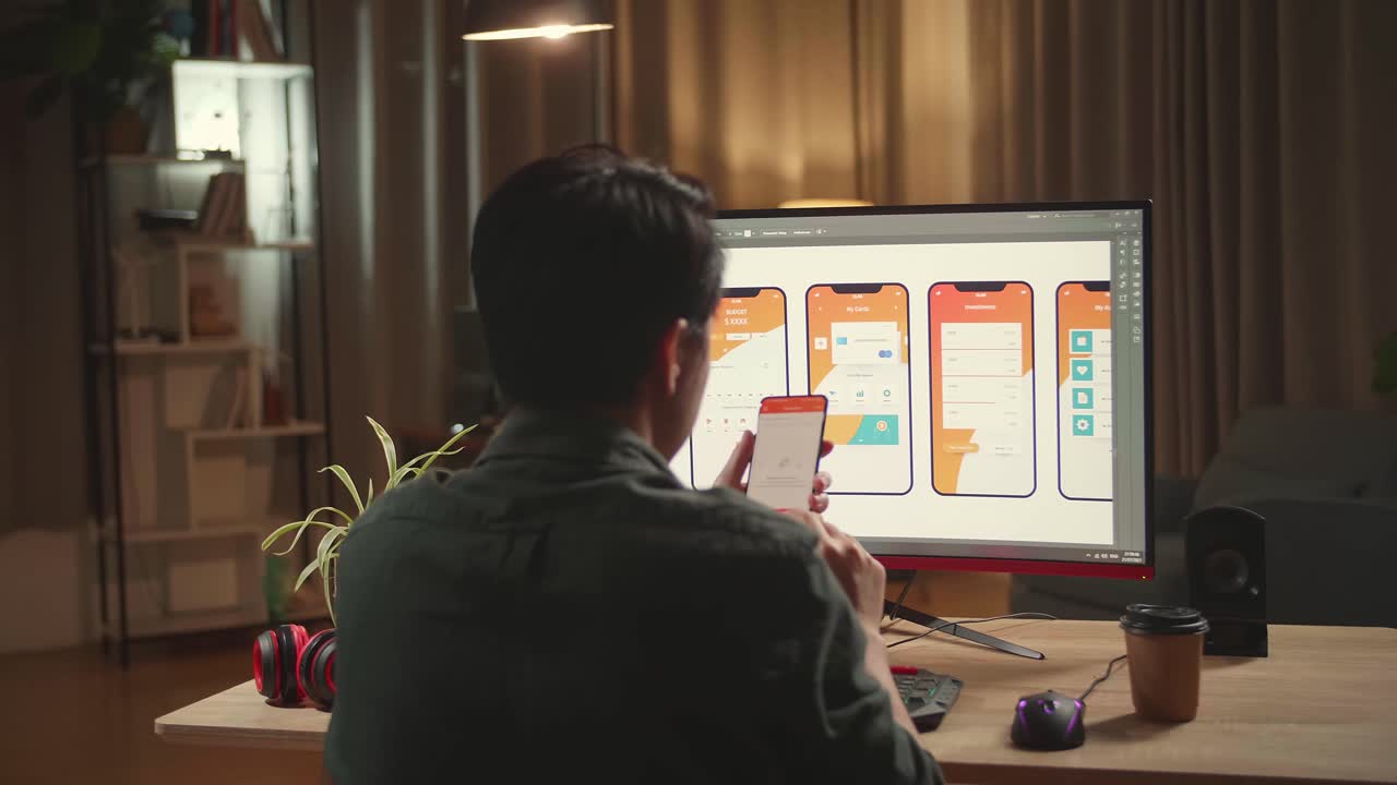 Asian Male Mobile Application Developer Works And Using Mobile Phone With Graphics On His Personal Computer. He Works At Night