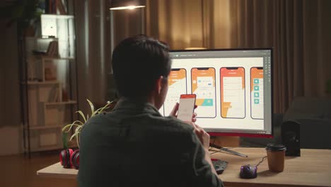 Asian Male Mobile Application Developer Works And Using Mobile Phone With Graphics On His Personal Computer. He Works At Night