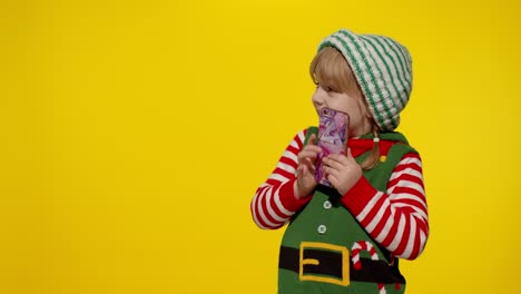 Kid-girl-with-mobile-phone-in-Christmas-elf-Santa-helper-costume-points-fingers-aside-on-blank-wall