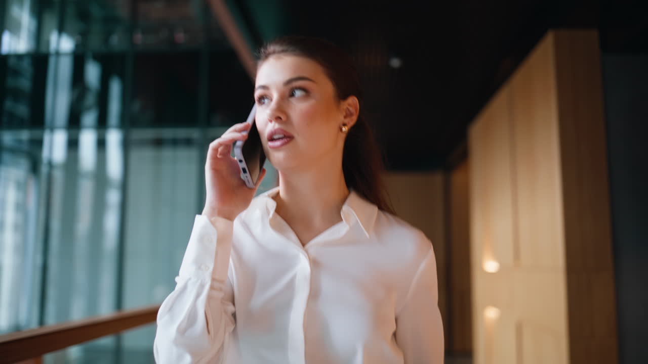Joyful brunette speaking smartphone walking business center hallway closeup