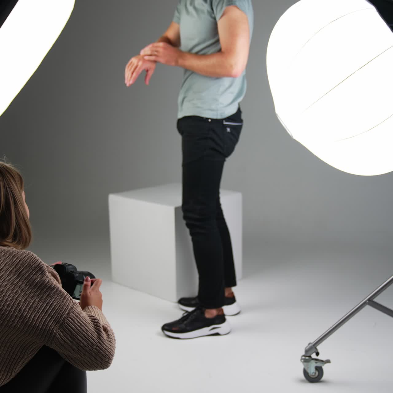 Male model posing for a photographer in studio. Woman taking pictures of man's footwear on white backdrop