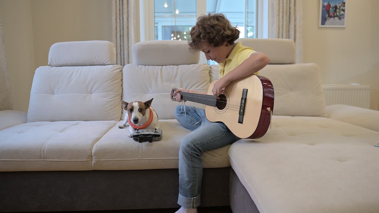 Blond boy with curly hair playing the guitar sitting on the couch, next to him is his dog lying