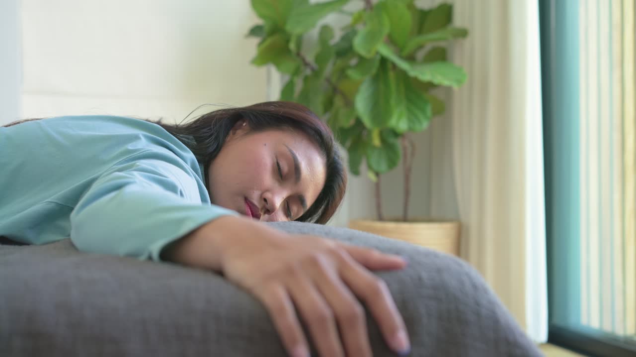 Cahaya yang menembus tirai membangunkan wanita Asia di sofa abu-abu yang mengulurkan tangan di atas bantal, ruang salin