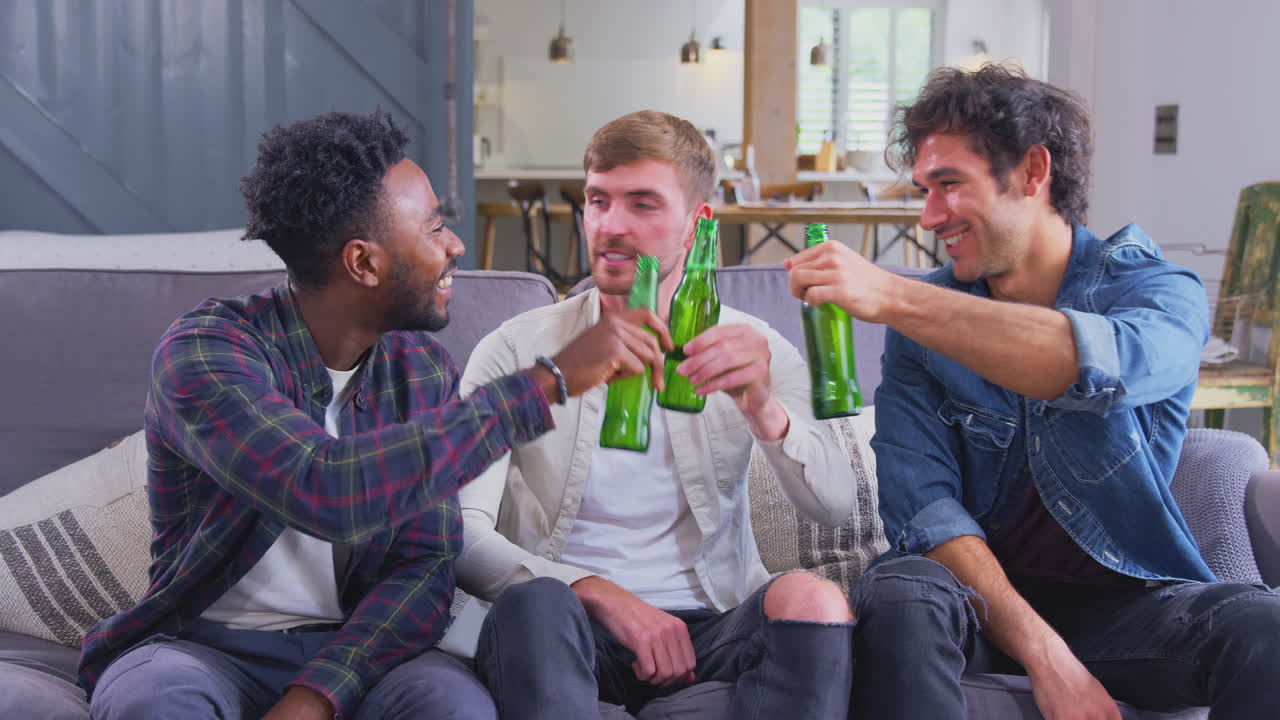 Multi-Cultural Group Of Male Friends On Sofa At Home Drinking Beer And Doing Cheers With Bottles