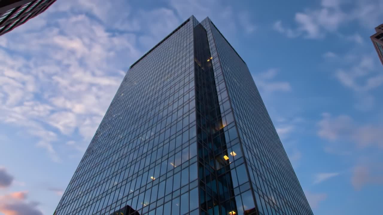 A tall glass building stands in the heart of a city, mirroring the colorful clouds of the evening sky. The architecture showcases a sleek design, while the lights start to illuminate inside.