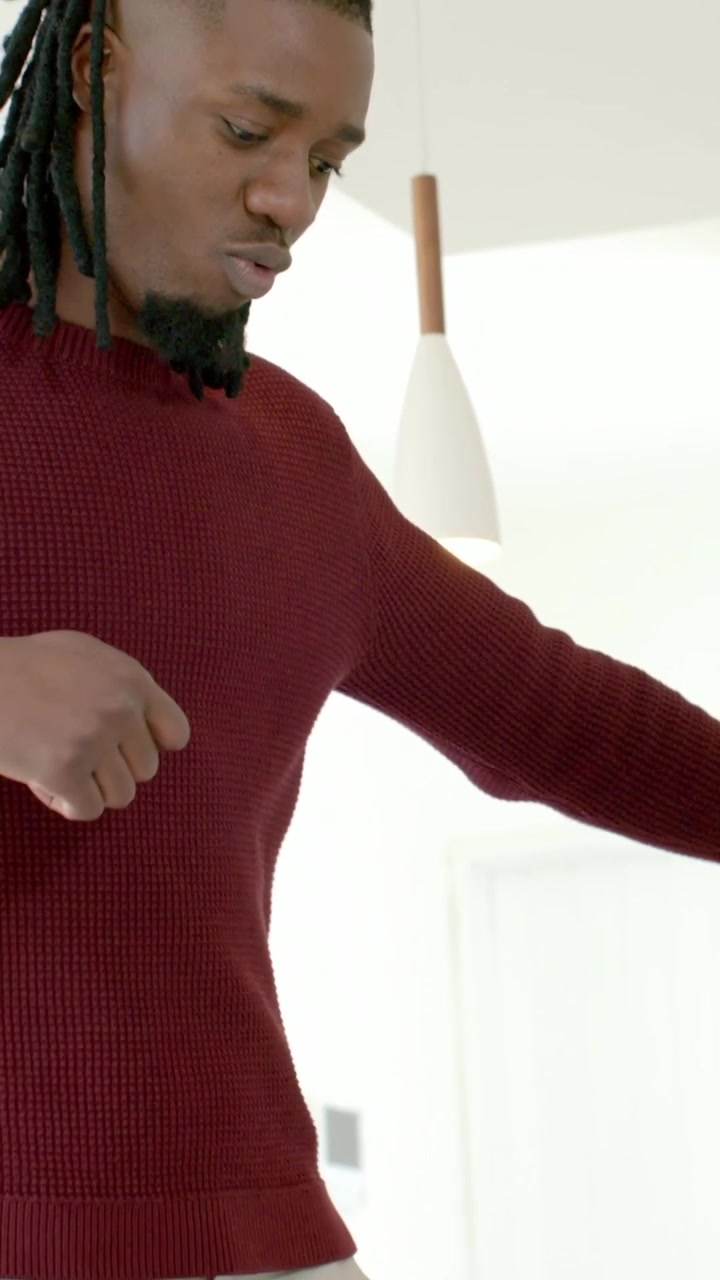 Vertical video: Filming man with dreads adjusting maroon sweater and beige cargo pants in kitchen