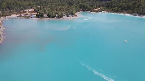 una toma aérea dinámica desde una gran altura de la bahía que se mueve hacia abajo pasando una moto de agua y yendo hacia la orilla del mar mientras sigue a los wakeboarders remolcados por un bote rápido