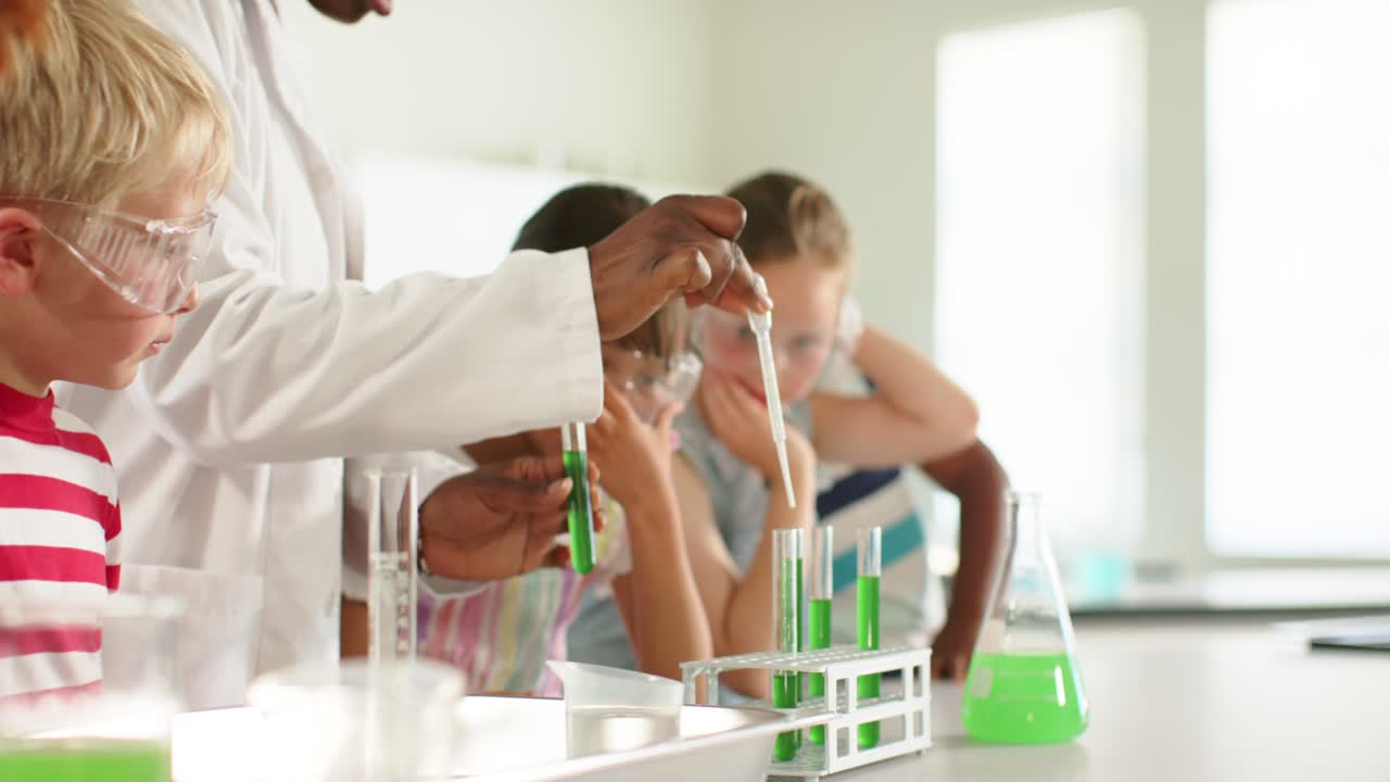 Male instructor in labcoat teaching kids with goggles dipping pipet into flask filling tubes in lab